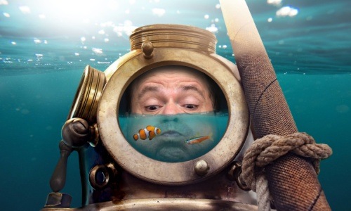 A photo of a man wearing a old fashioned diving helmet partially filled with water and 2 colorful tropical fish